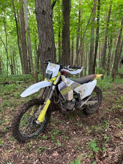 The image depicts a dirt bike lying on its side in a wooded area. The bike is white and yellow, with muddy tires and body. The background consists of trees and ground vegetation. The location, Gruzinskii, Russia, cannot be determined from the image. No people are visible.