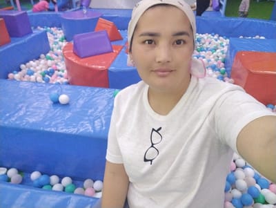 A young woman with light brown skin and dark hair, wearing a white t-shirt and a light-colored headscarf, is captured in a selfie. She is positioned in the foreground, with a blurred background of a colorful indoor playground. The playground features blue, red, and purple soft play structures and a ball pit filled with an assortment of white, blue, pink, and green plastic balls. In the background, two children can be seen, one wearing a pink shirt and the other a striped shirt. The overall scene suggests a recreational setting. The image does not contain specific landmarks that would allow for identification of the city of Talas City, Kyrgyzstan, or any other specific urban location.