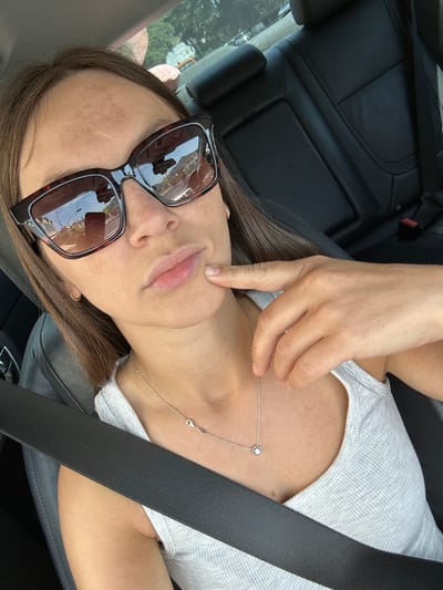 A close-up, waist-up shot shows a woman sitting in the driver's seat of a car, facing forward. She is wearing large, dark-rimmed sunglasses with reflective lenses, a light gray ribbed tank top, and a delicate silver necklace with a small, round pendant. Her right hand is raised, with her index finger touching her chin. Her brown hair is visible, framing her face. The interior of the car is visible, including black leather seats, a seatbelt across her chest, and the car's rearview mirror in the sunglasses' reflection. The background, visible through the car's windows, shows a blurred outdoor scene with trees and buildings, suggesting an urban or suburban environment. The woman is in Stanišići, Montenegro.