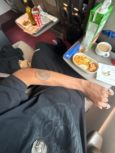 The image shows an individual sitting in an airline seat. A tray table is deployed with a half-eaten meal in a white dish, a white coffee cup, a juice box, a blue packet, and some jewelry on a white napkin. A second tray table with a partially consumed meal, beverage, and wine bottle is on the left. The person's arm, with a tattoo, is visible. The person wears a black shirt with a silver brooch. This image could be taken on a flight to or from Fretin, France, as Fretin is home to Lille Airport.