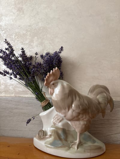A light brown porcelain rooster figurine stands on a light beige and blue base, placed on a wooden surface. Next to the rooster, a bunch of dried lavender is tied with twine and is held in a white ceramic vase. The background is a textured light beige wall. The scene is indoors, likely in a residential setting.