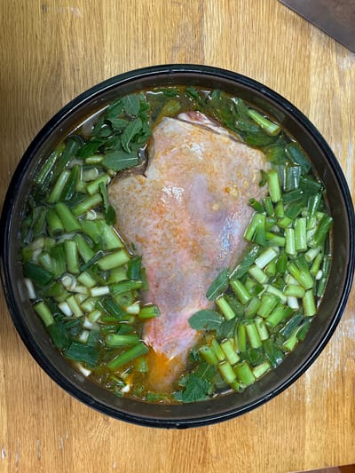 A top-down view shows a dark, circular bowl filled with a broth-like liquid, containing a large cut of raw meat, possibly pork or poultry. The meat is pale in color with some reddish-orange seasoning on its surface. Chopped green onions and mint leaves are scattered throughout the broth. The bowl sits on a light brown wooden surface. There is no evidence indicating the location is Oswestry, United Kingdom.