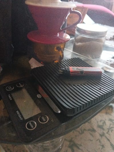 An indoor scene in Dogsenang Gorphoog, Bhutan, displays objects arranged on a patterned surface. A red coffee dripper with a white inner filter sits atop an amber-colored glass mug in the upper left quadrant. Below, a black digital scale, featuring a blank display and buttons labeled "TIMER," "WEIGHT," "TARE," "ON/OFF," and "MODE," is partially visible. A black rectangular rubber mat with prominent horizontal ribs rests on the scale's platform. Positioned horizontally on this mat is a red and black disposable cigarette lighter, which bears a white label on its side. To the right, a clear glass jar with a metallic lid contains a light brown granular substance. The overall arrangement suggests a coffee preparation area.