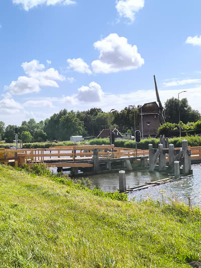 In Koedijk, Netherlands, a wooden bridge spans a body of water under a bright blue sky with scattered white clouds. The bridge has a light brown wooden railing and appears to be a drawbridge or lift bridge, with sections of wooden planks extending into the water. Several wooden posts are visible along the water's edge, some of which are part of the bridge's mechanism. To the right of the bridge, a traditional Dutch windmill with a thatched roof and wooden sails stands prominently. The windmill is surrounded by lush green trees and vegetation. In the background, there are a few houses and buildings, and a traffic light with a red signal is visible near the bridge. The water is calm with reflections of the sky and surrounding elements. The foreground is dominated by a grassy embankment.