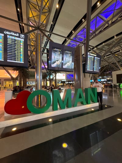 Inside an airport terminal, the large letters "OMAN" stand vertically. A red heart replaces the base of the "O." Above the letters, a person wearing a white shirt stands. Departure and arrival information screens flank the letters, displaying schedules. The background shows the airport's interior, with support structures, palm trees, shops, and people milling around. The floor has a polished, dark surface with embedded lights.