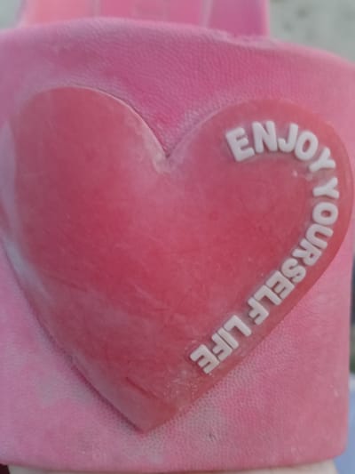 The image is a close-up shot of a pink heart-shaped object with white text on it. The text reads "ENJOY YOURSELF LIFE" and is arranged in a curve around the upper and right edges of the heart. The heart appears to be made of a soft, possibly rubber or silicone material, and is attached to a textured pink fabric. The background is out of focus but suggests a pink, possibly fuzzy surface. The scene is well-lit, and the focus is sharp on the heart and text. There is no discernible context of a city or specific location like Altinkul, Uzbekistan within the image itself.