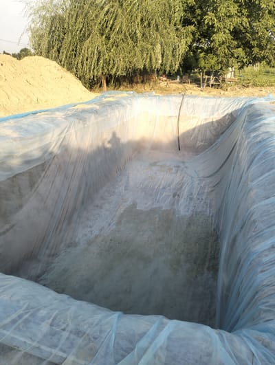The image shows an empty, rectangular pit lined with a translucent blue plastic tarp. The tarp is draped over the sides of the pit and extends outwards, creating a temporary covering. The bottom of the pit appears to be rough and uneven, with some debris and dirt visible. In the background, there is a large pile of sand or dirt to the left, and several trees with lush green foliage to the right. The overall scene suggests an outdoor construction or excavation site in a rural or semi-rural area. The mention of Aktam (Tashminvody), Uzbekistan, indicates a specific geographical location, which could be relevant for context regarding the climate, vegetation, and typical building practices. The lighting suggests it is daytime, with sunlight casting shadows across the plastic tarp. There are no people clearly visible in the image, though a faint shadow on the tarp might indicate a person's presence at some point. The environment is natural, with soil, sand, and trees dominating the landscape.