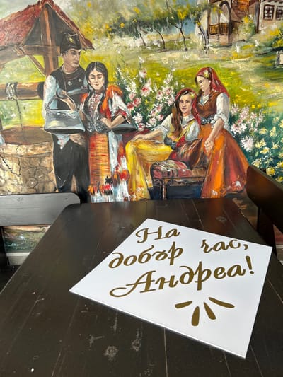 A close-up, slightly angled shot captures a white sign with golden Cyrillic lettering placed on a dark wooden table. The sign reads "На добър час, Андреа!" which translates to "Good luck, Andrea!". Behind the table, a vibrant, slightly worn mural depicts a pastoral scene with figures in traditional Bulgarian attire, possibly from Ruska Bela, Bulgaria, set against a backdrop of rustic buildings and lush greenery with flowers. The mural shows four people: a man and a woman near a well on the left, and two women seated among flowers on the right. The overall impression is of a cultural or historical setting, possibly a restaurant or a community space.