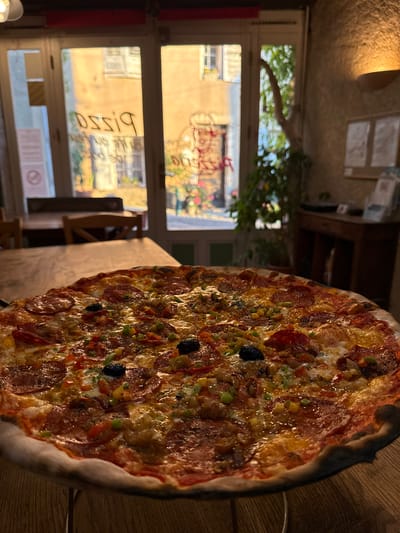 A close-up, eye-level shot showcases a large, round pizza topped with pepperoni, olives, bell peppers, and corn, resting on a wooden table. In the background, large glass doors reveal a sunlit street scene in Moustiers-Sainte-Marie, France, with buildings adorned with flower boxes and shops displaying signage, including the word "PIZZA" in reverse. The warm, natural light spills into the indoor space, illuminating a dimly lit dining area with wooden furniture and a warm-toned wall. The overall impression is that of a cozy pizzeria on a bright day.