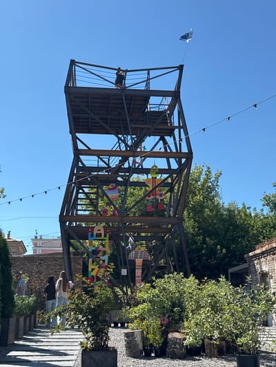 An outdoor scene features a tall, metal, lattice-like structure with multiple platforms and stairs. A flag flies from the top. Colorful, cartoonish figures are attached to the structure at various levels. In the foreground, people walk along a path lined with greenery in planters and decorative logs. The background shows brick buildings and trees under a clear blue sky. The location is identified as a park in the city of Yelets, Russia.