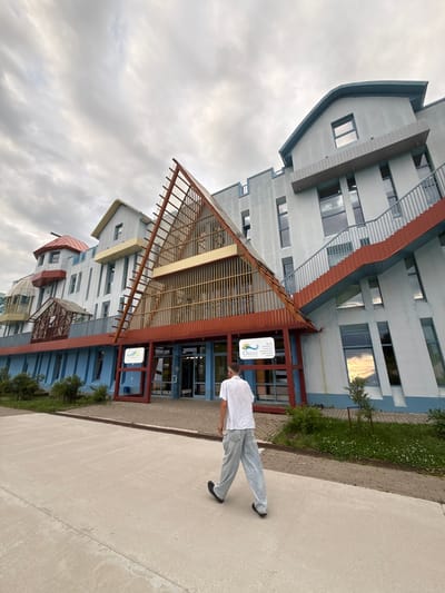 In Petrovo, Russia, a person walks away from the camera on a paved pathway in front of a distinctive, multi-story building with a light blue facade and red architectural accents. The building features a large, triangular, glass-enclosed structure with wooden beams that extends from the main facade, and a prominent staircase with red risers and blue railings on the right side. The sky is overcast with white clouds. Greenery and bushes are visible along the base of the building. A sign with Russian text and a logo is mounted near the entrance.