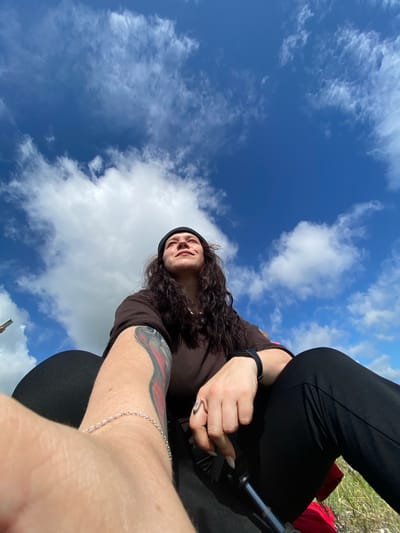 In an outdoor, ground-level, wide-angle selfie, a person with long, dark, wavy hair, wearing a black beanie, a brown long-sleeved shirt, and black pants, is captured. The person is looking upwards, with their face illuminated by sunlight. A tattoo is visible on their left arm, and they are wearing a silver bracelet on their left wrist and a black smartwatch on their right wrist. Their left hand rests on their left knee, and their right hand is positioned near their right knee. The background is dominated by a bright blue sky with scattered white clouds. The location is identified as Andreevka, Russia. The foreground shows a hint of grass and what appears to be an object, possibly a camera or phone, in the lower left corner, indicating the selfie was taken by the subject.