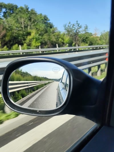 The image captures a view from inside a moving vehicle, looking into the side-view mirror. The mirror reflects a highway scene with a clear blue sky above. The highway has a guardrail running alongside it, and trees and greenery line the sides of the road. In the distance, other vehicles can be seen on the highway. The foreground shows a portion of the car's interior, including the side mirror housing. The location is identified as Novi han, Bulgaria.