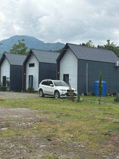 This image depicts a row of three modern, minimalist cabins situated in Kapreshumi, Georgia. The cabins feature a dark grey, pitched roof and a contrasting white and dark grey facade. Each cabin has a single door and a small window. In front of the cabins, a white SUV is parked on a gravel and grass lot. Behind the cabins, a lush green mountain range is visible under a cloudy sky. To the right of the cabins, a large blue water tank and a tall, slender evergreen tree are present. The overall scene suggests a tranquil, natural setting with contemporary accommodations.