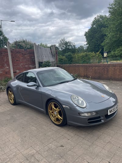A grey Porsche 911 is parked on a paved driveway next to a brick wall. The car has gold alloy wheels and a black license plate that reads "NK55 HUL". In the background, there are trees and a wooden fence. The sky is cloudy, suggesting it may be an overcast day. The scene is set in Wolverhampton, United Kingdom. There are no people visible in the image.