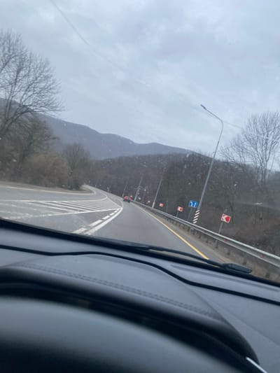 A roadside view from inside a vehicle shows a gray, overcast day. The road curves ahead, lined with white stripes, yellow painted lines, and guardrails. Roadside signs and streetlights dot the landscape. A tree line and distant mountains fade into the misty background. The windshield of the vehicle shows a slight streak, indicating moisture or imperfection.