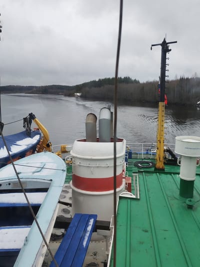 The photograph shows a view from the deck of a vessel on a body of water, likely in Depo, Russia, given the text on the white container, which is in Russian. The deck is green and features various equipment, including a red and white stack, two lifeboats covered in snow, and a yellow crane. A tall, black pole with a ladder and some attachments rises from the deck.
In the background, a river or lake is surrounded by trees with sparse patches of snow on the ground. The sky is overcast, and the overall light suggests a cold or wintry day. There are no people visible in the scene. The photograph appears to be taken from an elevated position on the ship, providing a panoramic view of the landscape.