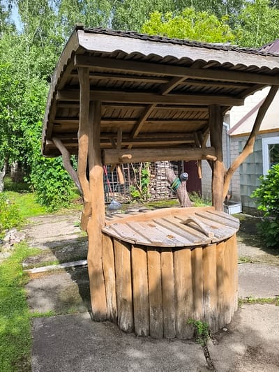 This is a medium shot, eye-level, outdoor photograph of a rustic wooden well structure located in the village of Žiaukos, Lithuania. The well is made of vertical wooden planks for the barrel and a wooden roof supported by rough-hewn wooden posts. The roof is covered with dark shingles. The well is situated on a concrete patio, with grass and a dirt path surrounding it. In the background, there is lush green foliage, trees, and a building with a light-colored brick wall and a window. A garden rake is leaning against the well structure. There are no people visible in the image. The lighting suggests it is a sunny day.