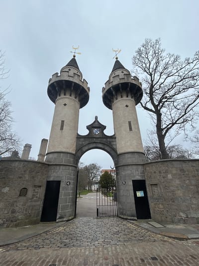 The image shows the Brig o' Balgownie, a medieval bridge located in Aberdeen, Scotland, United Kingdom. The bridge's towers are visible, featuring conical spires topped with weathervanes. The towers are constructed of gray stone and are adorned with decorative elements. An arched opening in the center allows passage through the structure, revealing a view of a street with a barrier. The area surrounding the bridge is paved with cobblestones. A bare tree branch appears in the upper portion of the image, and the sky is overcast. Number plaques "41" and "51" are seen.