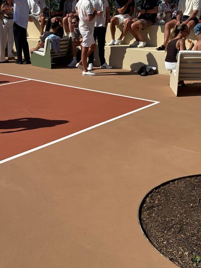 An outdoor gathering on a brown surface marked with white triangle patterns. People, including seated individuals, are situated along the edge. To the right, there's a circular patch filled with dark soil and possibly foliage. The scene is brightly lit, casting distinct shadows of people onto the surface.