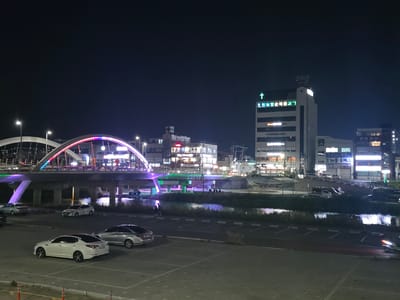 This is an eye-level, medium shot of a streetscape at night in Eumnae-ri, South Korea. In the foreground, four cars are parked in a lot: a white Kia Optima, a silver Hyundai Sonata, a silver sedan, and another silver sedan. The lot is paved with gray interlocking bricks.
In the midground, a river or canal runs horizontally across the frame, reflecting the lights from the surrounding cityscape. On the far bank of the water, a grassy area with some trees is visible, bordering a raised concrete walkway. Further beyond the walkway, a bridge arches over the water, illuminated with colorful lights in shades of purple, pink, and green. Several people are visible on and under the bridge, and some are gathered by the riverbank.
The background of the image features a collection of buildings. Prominently, a tall, multi-story building with a green cross symbol and Korean text reading "조천제일순복음교회" (Jocheon Jeil Soonbokum Church) stands out on the right side of the frame. Other buildings of varying heights, some with illuminated windows and signage, line the street. The sky is dark, suggesting nighttime.