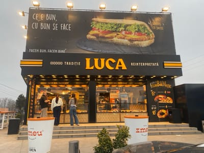 The image displays the exterior of a LUCA establishment in Agigea, Romania, under an overcast sky. The building features a black facade with the name "LUCA" in large orange letters and the slogan "TRADIȚIE REINTERPRETATĂ" (Tradition Reinterpreted). Above the structure, a large billboard is illuminated by multiple overhead lights, showcasing a "SANDVIȘ CU ȘNIȚEL" (Schnitzel sandwich) and text in Romanian, including "CE E BUN, CU BUN SE FACE" (What is good, is made with good) and "FACEM BUN. FACEM BINE." (We do good. We do well/good).

The ground level of the establishment consists of large glass display windows revealing an interior with numerous baked goods and counter areas. Three individuals are visible inside: one male, wearing a dark shirt and light pants, interacting with a female wearing a light-colored top and dark pants. A second female, wearing a striped top and blue jeans, stands further to the right, facing a display. Steps lead up to the entrance.

In front of the building, two large, white, coffee cup-shaped containers are present, labeled "Cafea zmeu artizanală" (Zmeu artisanal coffee) and "oferită de LUCA" (offered by LUCA). To the right of the entrance, a black outdoor menu board with white and red text advertises various food combinations and prices, such as a "Combunație" (combo) for 13 ROL. Bare trees and small green bushes are visible to the left and directly in front of the building. The partial hood and windshield reflection of a dark vehicle are visible in the lower right foreground.