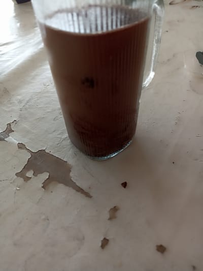 This is a close-up, slightly blurry, eye-level shot of a glass mug filled with a dark, opaque liquid, likely hot chocolate or coffee, placed on a table covered with a patterned, light-colored plastic or vinyl tablecloth. The mug is clear glass with vertical ribbed texture and has a prominent handle. The liquid fills most of the mug, with a small air gap at the top.
The tablecloth shows signs of wear, with peeling patches revealing a darker surface underneath, and several dark stains, possibly from spills, are visible on it. One larger, irregular stain is prominent to the left of the mug, and smaller dark specks are scattered across the surface. The lighting suggests an indoor setting with diffused natural light, possibly daytime, but no specific time or weather conditions are discernible. There are no people visible, and the focus is entirely on the mug and the tabletop. No text is visible in the image. The overall impression is a casual, everyday moment.