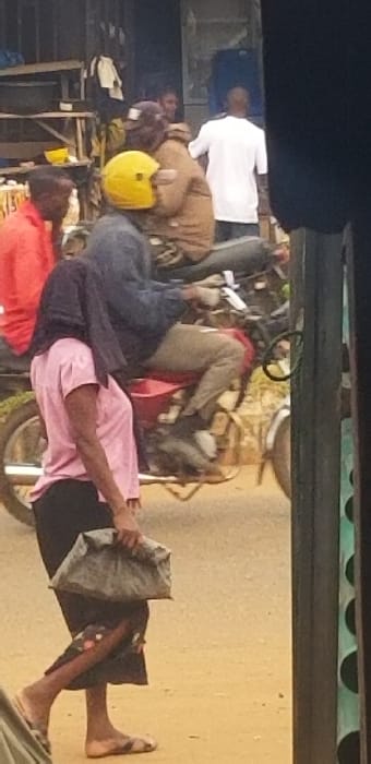 The image depicts an active street scene in Ado-Ekiti, Nigeria. In the foreground, a woman of African descent walks from left to right, wearing a light pink short-sleeved top, a dark patterned skirt, and light-colored flip-flops. Her head is covered by a dark fabric. She carries a grey, irregularly shaped bundle in her right hand. Directly behind her, a red motorcycle is visible, carrying a driver and a passenger. The driver wears a dark blue jacket, light-colored pants, and a prominent bright yellow helmet. The passenger, seated behind the driver, wears a brown jacket and dark headwear. In the mid-ground and background, several other individuals are present. To the left, a man in a red shirt is partially visible. Further back, a person in a white t-shirt stands facing away from the camera. The environment includes structures resembling market stalls or shops with wooden framing, some displaying hanging items. The ground is a light, unpaved surface. A dark, vertical pole with green circular accents frames the scene on the far right.
