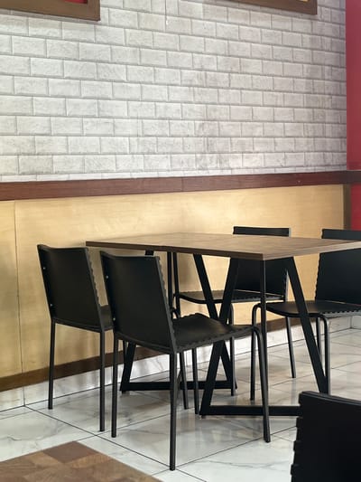 The image depicts an indoor dining area with a light-colored brick-patterned wall and a wooden paneled lower wall. A rectangular wooden table with black metal legs is centrally positioned, surrounded by four black chairs. The floor is tiled with a marbled pattern. To the right of the scene, a red wall is visible. The lighting suggests an indoor environment, possibly a restaurant or cafe in Meteseh, Indonesia. There are no people present in the image.