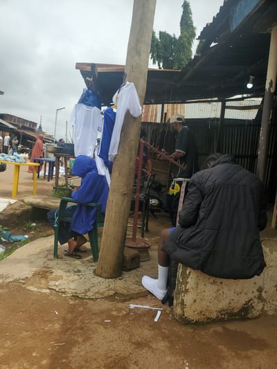 The image depicts a market scene in Ohit, Nigeria. In the foreground, a person wearing a blue hooded sweatshirt and dark shorts is seated on a green plastic chair, looking down at their phone. To the right, another person, seen from the back and wearing a black puffer jacket and white socks with sneakers, sits on a concrete block. Behind them, under a corrugated metal roof structure, two other individuals are visible. One person, wearing a cap and a dark t-shirt with white trim, appears to be engaged in some activity, possibly weaving or crafting, holding a wooden object with multiple prongs. Another person is partially visible in red attire to the left. Several white and blue garments, possibly clothing for sale, are hanging on a wooden pole in the middle of the scene. A yellow table with items on it is situated to the left, with other market stalls and structures visible in the background. The ground is a mix of dirt and broken concrete. The sky is overcast.