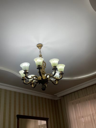 The image shows an ornate chandelier hanging from a white ceiling. The chandelier has multiple arms and pale green shades. The ceiling has a curved cove detail. To the right of the chandelier is a wall with striped wallpaper and a framed picture. The overall environment appears to be the interior of a building. The location is not discernible from the image.