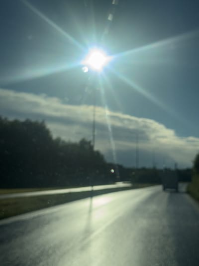The image depicts a sun-drenched, blurred road scene in Harkujärve, Estonia. The bright sun, positioned in the upper portion of the frame, creates a strong lens flare that washes out much of the sky and the road ahead. The road appears to be wet, reflecting the sunlight and giving it a shimmering appearance. On the left side of the road, a dark mass of trees or foliage is visible, also blurred due to the focus. In the distance, a faint silhouette of a vehicle is present on the right side of the road. The overall impression is one of driving during a bright, possibly humid, day. The extreme blurriness and lens flare suggest the photo was taken from a moving vehicle, likely through a window. There are no discernible people or specific landmarks to identify a precise location within Harkujärve.