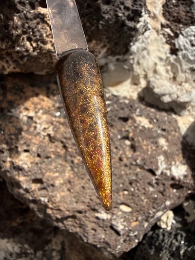 The image shows a sample of brown and gold glitter nail polish held up against a background of volcanic rock. The setting is likely in Yaiza, Spain, given its volcanic landscape. The nail polish sample is elongated and tapers to a point. It is affixed to a clear, flat holder. The volcanic rocks in the background are porous and display a range of brown and grey tones. Some white mineral deposits are also visible on the rocks. The image is well-lit, with the sunlight highlighting the glitter in the nail polish.
