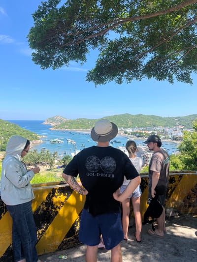 The image shows a group of four individuals standing on an elevated viewpoint overlooking a bay in Vĩnh Hải, Vietnam. The viewpoint has a yellow and black striped barrier. The individuals are positioned from left to right: a person wearing a light-colored hooded jacket and jeans, looking down at something in their hands; a person in the center wearing a black t-shirt with a graphic design and blue shorts, with their hands on their hips; a person behind them wearing a white t-shirt and denim shorts, facing away from the camera; and a person on the far right wearing a grey t-shirt and dark shorts, also facing away from the camera.
The bay below is filled with numerous small boats. In the middle ground, there is a village with buildings and more boats. The background features lush green hills and a clear blue sky with some light clouds. The foreground is dominated by the viewpoint and the people. The overall scene suggests a scenic overlook in a coastal Vietnamese town.