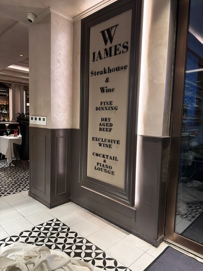 A ground-level close-up shot of a signage for "W James Steakhouse & Wine" in Qinhuai District, China. The sign is dark grey with light text detailing the restaurant’s offerings, including fine dining, dry-aged beef, exclusive wine, and a cocktail & piano lounge. The wall supporting the sign is beige with a textured finish. The scene includes a glimpse of the establishment's interior through the window at the top, and the floor is tiled in a black and white pattern. Parts of tables, chairs, and decor inside the restaurant can be seen through the doorway.