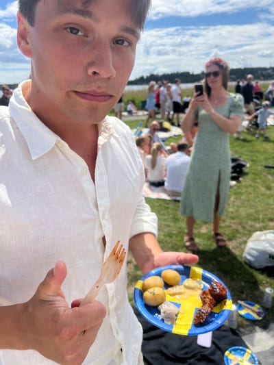 In Österskär, Sweden, a man in a white linen shirt is holding a plate of food with a wooden fork. The plate is decorated with the Swedish flag. On the plate are small potatoes, some type of fish or seafood in a creamy sauce, and what appears to be grilled meat. The man is looking towards the camera, and his thumb is extended.

In the background, a gathering of people is enjoying a sunny day outdoors. A woman in a green floral dress with a slit and a flower crown is holding a smartphone, possibly taking a picture or video. Other people are sitting on blankets on the grass, and some are standing and interacting. The environment suggests a picnic or outdoor celebration. Swedish flags are visible on other plates and scattered around the area, indicating a national celebration, likely Midsummer given the setting and attire. The presence of a body of water in the distance suggests the location is near the coast.