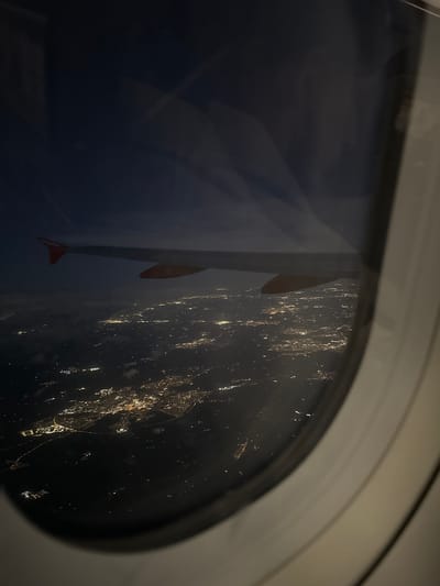 The image shows a view from an airplane window at night. The wing of the plane, painted with the "easyJet" logo, is visible. Outside the window, city lights are seen scattered across the dark landscape. It is not possible to ascertain a specific location in the city of Stafford, United Kingdom from the available image.