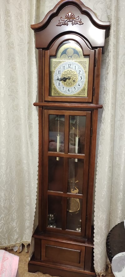 This media file is a full-shot, vertically oriented image of a grandfather clock. The clock is made of dark wood and features a decorative top with carved detailing. Below the top, a glass-enclosed section reveals the clock's face and pendulum. The clock face is round with Roman numerals and ornate gold detailing. The hour and minute hands are black and appear to be set at approximately 9:12. Above the main clock face is a smaller dial, likely for moon phases or other complications, with a blue background and celestial imagery.
The clock has a glass-doored cabinet in the lower section, revealing hanging weights and the pendulum. The pendulum itself is golden and has a decorative disc at its base, showing intricate carvings. Visible within the cabinet are some decorative objects, appearing to be small metallic figurines and a larger round decorative element.
The grandfather clock is positioned indoors, in what appears to be a living room or hallway. It is set against a backdrop of cream-colored curtains with a subtle, ornate floral pattern. The lighting suggests it is daytime, but the weather is not discernible. There is no visible text in the image. To the left of the clock, a pink cloth is partially visible on the floor, and to the right, a small portion of a round, dark wooden table with carved edges is seen. The overall impression is of a well-maintained, traditional piece of furniture in a domestic setting.
