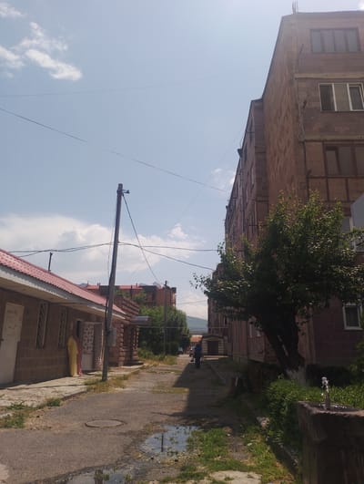 The image depicts a street scene in Nor Hachin, Armenia. In the foreground, a paved road with puddles of water leads towards the center of the image. To the left, a low, single-story building with a red tiled roof and brick walls is visible, featuring several windows and doors. Two figures, one in a yellow garment and another in a pink garment, are standing near the entrance of this building. Adjacent to this building, a utility pole with wires extends upwards.

Dominating the right side of the image is a multi-story brick building, likely an apartment complex, with multiple windows. A tree with green foliage stands in front of this building, its trunk painted white at the base. Further down the street, a person in a blue shirt and dark pants is walking away from the camera, carrying bags. In the background, other buildings and trees are visible, and a mountain range can be seen in the distance. The sky is partly cloudy. A stone fountain with water spouting is positioned to the right of the paved path. The overall impression is that of a residential area in a town or city.