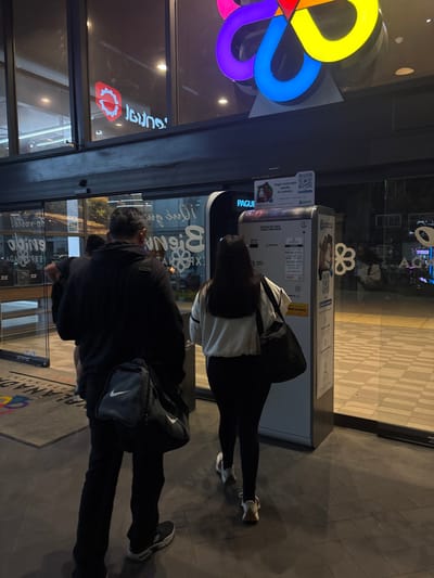Two people are interacting with a payment kiosk outside a building with glass walls. One person, wearing a black jacket and pants with a Nike crossbody bag, stands slightly in front. The other person, wearing a white top and black leggings, is closer to the kiosk, appearing to be making a transaction. The building has colorful logo displays and signage visible through the glass. The ground is paved and leads into the building.