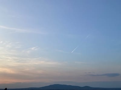 The image captures a wide shot of the sky over Arandjelovac Municipality, Serbia, likely during sunset or sunrise. The sky is a gradient of colors, transitioning from a soft pink and orange hue near the horizon to a clear blue higher up. Thin, wispy clouds are scattered across the sky, with two distinct contrails visible, suggesting aircraft have recently passed through. In the foreground, a silhouetted mountain range stretches across the landscape, with faint indications of structures or terrain at its base. The overall atmosphere is serene and expansive, emphasizing the natural beauty of the sky and the surrounding environment.