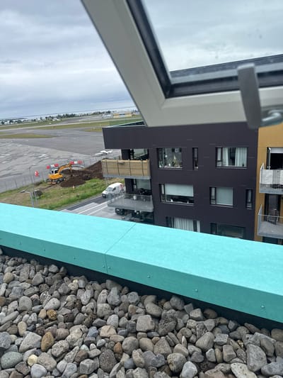 The image captures an outdoor scene from an elevated perspective, likely from a rooftop or balcony, overlooking an airport in Reykjavik, Iceland. In the foreground, a turquoise-colored barrier lines the edge, with a bed of grey and brown river rocks beneath it. Beyond this, a modern, dark-colored apartment building with multiple windows and balconies is visible. Adjacent to it, a lighter-colored building with wooden paneling stands.
Further into the scene, the grounds of an airport are spread out. A yellow excavator is actively digging a mound of earth, surrounded by temporary fencing and traffic cones, suggesting ongoing construction or maintenance work. Several small airplanes are parked on the tarmac. The sky is overcast, with a vast expanse of grey clouds. In the distance, a body of water and a treeline can be observed, characteristic of Reykjavik's coastal landscape. A white van is parked on a road near the construction site, and a black car is visible on a lower level of the apartment building.