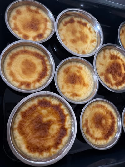 A high-angle, medium shot captures a tray of crème brûlée desserts arranged in a black, oven-like environment. The desserts are presented in individual, round, silver ramekins, with eight fully visible and one partially visible at the top right. Each dessert has a golden-brown, caramelized sugar topping that exhibits varying degrees of browning and crackling. The creamy custard beneath the caramelized layer is a pale yellow. The lighting suggests a flash or overhead light source, creating reflections on the metallic ramekins. The scene is sterile and focused solely on the food presentation. There is no indication of a specific location within Zmeitsa, Bulgaria, beyond the implied setting of a kitchen or a place where baked goods are prepared or served. The image contains no people or any other objects.