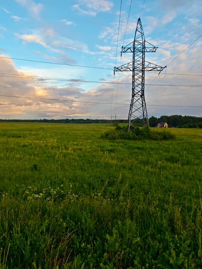 A low-angle, eye-level photograph depicts a grassy field under a partly cloudy sky, with a power transmission tower as the dominant feature. The tower, constructed of dark gray metal, stands prominently in the middle ground, its open lattice structure extending high into the sky. Three power lines extend horizontally from the tower. The field is lush green, with varying textures and heights of grass. In the far distance, a few trees are visible. A yellow house is faintly visible in the background. The sky displays a mix of blue and white clouds, with hints of orange and pink near the horizon. The location is near Курово, Russia.