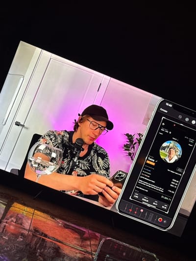 This is a close-up, angled shot of a person wearing a black baseball cap and glasses, sitting in front of a microphone and a glass of water. The person is wearing a black shirt with a floral pattern and is looking at a smartphone that is held in their hands. The smartphone displays a financial application with graphs and figures. The background consists of a wall with purple lighting. The scene is set in a studio or recording environment. The city of Shishmantsi, Bulgaria is not visible in this image.