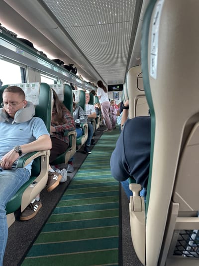 The image depicts the interior of a train carriage. In the foreground, a man wearing a light blue t-shirt and jeans is seated, leaning his head on a gray neck pillow. He has earbuds in and is wearing glasses and a wristwatch. To his right, a woman in a plaid shirt is seated, and further down the aisle, a man in a gray hoodie and jeans is visible. At the end of the carriage, a woman in a white shirt and pink pants and a young girl in a pink tracksuit are standing. Luggage is stored in the overhead compartments. The train has green and blue striped carpeting on the floor and green seats with light beige headrests and armrests. The ceiling features a patterned design with lights. The overall scene suggests a passenger train journey. The image was taken inside the Sobinka train station, located in Sobinka, Russia.