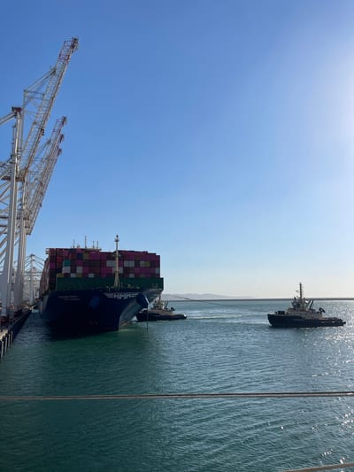 In the Moroccan city of Ksar al Majaz, a massive container ship, identified by the "HMM HANGADA" inscription, is seen maneuvering in a clear blue sky and calm, turquoise waters. The ship, laden with numerous colorful shipping containers, is docked alongside a port structure, with its bow facing the viewer. Two tugboats are actively assisting the large vessel, one positioned on its starboard side and another further out in the water, connected by a tow line. The port is equipped with tall, white gantry cranes, their structures stretching towards the sky. The background shows a hazy coastline with mountains in the distance. The sun is shining brightly, casting a glare on the water.