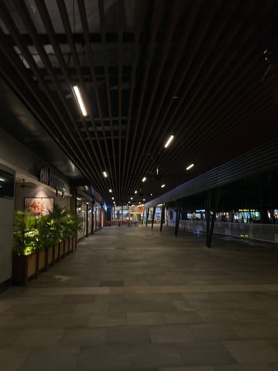The image depicts an outdoor pedestrian walkway at night in Putrajaya, Malaysia. The walkway is illuminated by integrated linear lights within a parallel-lined structural barrier. The walkway surface is paved with rectangular gray stone tiles. Towards the bottom of the frame, there is a landscaped area featuring tropical plants. An advertisement or poster is visible on the side wall. In the distance, faint lights suggest a commercial or entertainment area. The scene is generally devoid of people.