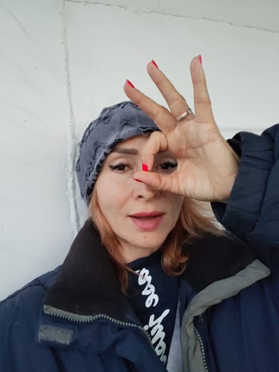 The image shows a medium close-up of a woman with light skin, blonde hair, and eye makeup. She is wearing a gray patterned bandana, a dark blue jacket with a black collar, and a dark t-shirt with white lettering. Her right hand is raised to her face, with her thumb and index finger forming a circle around her right eye. Her fingernails are painted bright red. She is looking directly at the camera with her mouth slightly open. The background is a textured white wall. The image does not contain any discernible features that would place it in Ivanov Bor, Russia.