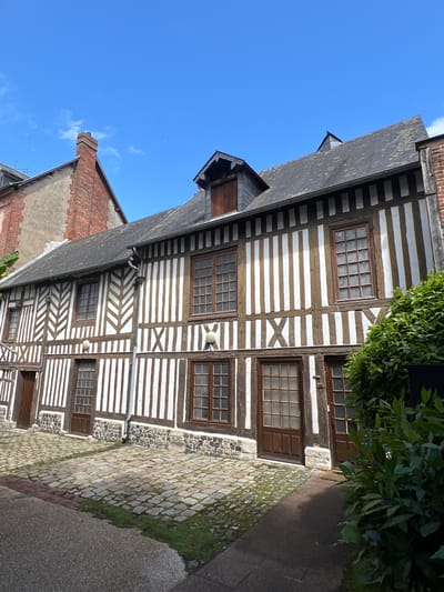 This is a medium shot, captured from a slightly low angle, of a traditional half-timbered building with a cobblestone courtyard in Honfleur, France. The building, predominantly white with dark wooden beams, features a steeply pitched slate roof with dormer windows. The facade is characterized by its intricate half-timbering, with vertical beams and diagonal bracing patterns. Several multi-paned wooden windows and doors are visible, some with stone sills. The cobblestone courtyard in front is uneven and interspersed with patches of grass. To the right of the building, dense green foliage partially obscures the view. In the background, a portion of another building with a red brick chimney is visible against a clear blue sky with a few wispy clouds. The scene appears tranquil, with no people or animals present.